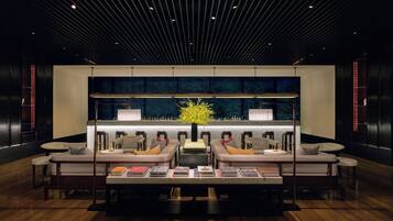 A modern lounge area with a central seating arrangement, a bar with a large window, and a wooden floor.
