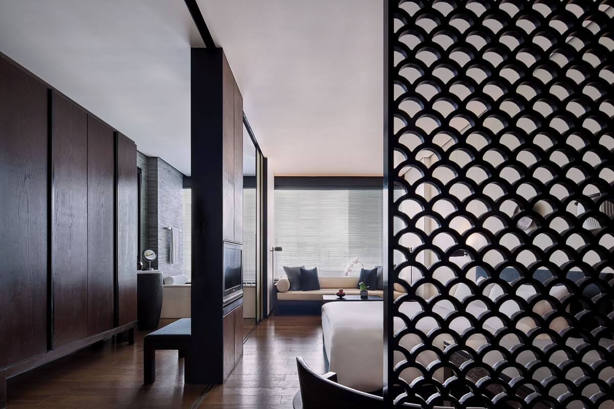 A modern hotel room with a large bed, a seating area with a sofa and pillows, a TV, and a decorative wall panel.