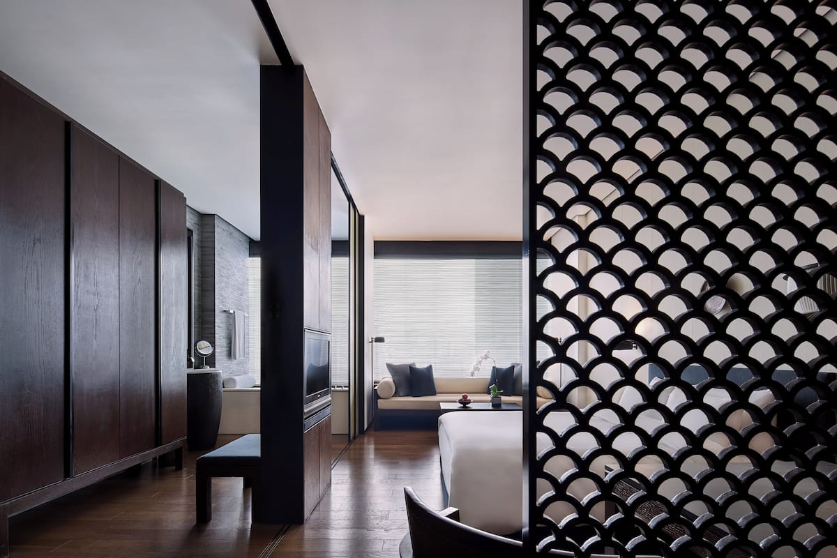 A modern hotel room with a large bed, a seating area with a sofa and pillows, a TV, and a decorative wall panel.