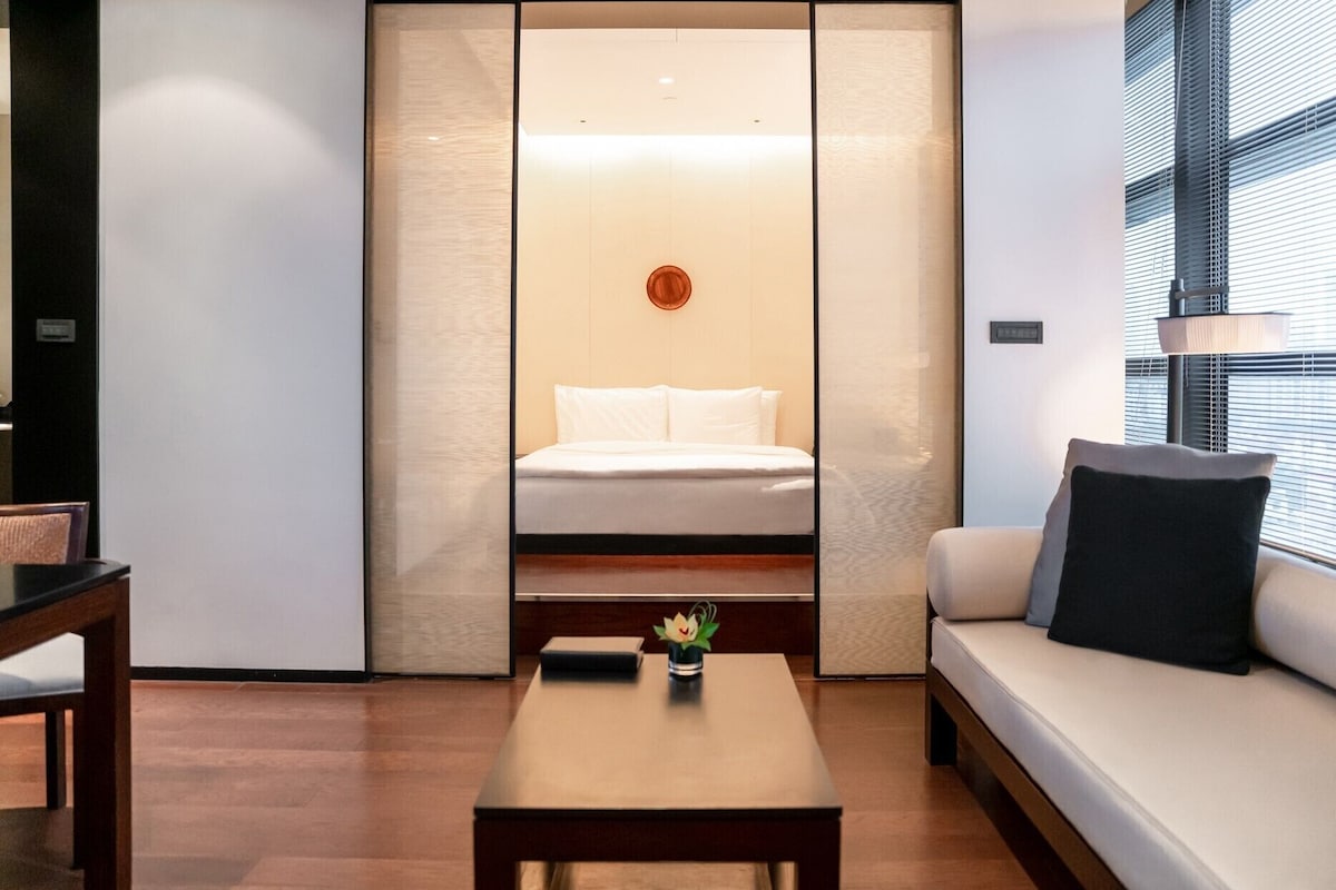 Une chambre d’hôtel moderne, équipée d’un lit, d’un canapé, d’une table basse et offrant une vue sur l’extérieur grâce à de grandes fenêtres.