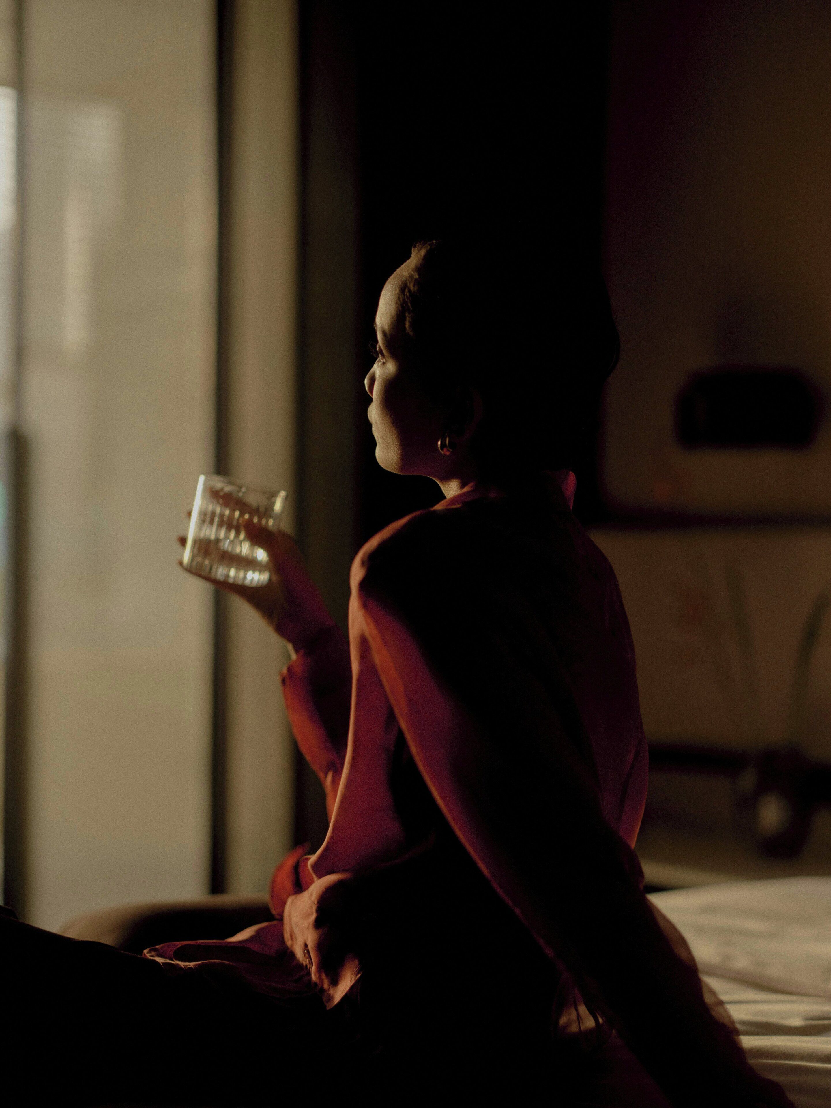 A person sitting on a bed with a book, in a dimly lit room with a window and a wall-mounted object.
