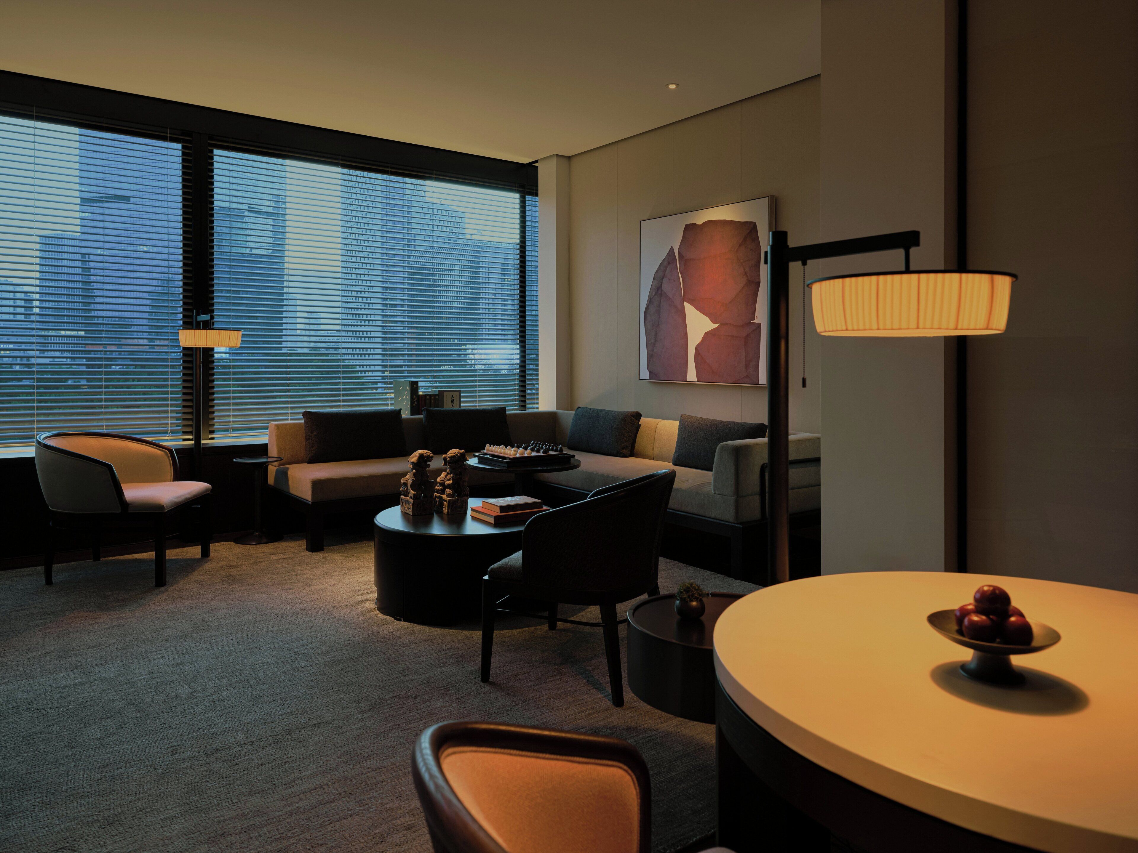 A modern hotel room with a sofa, armchairs, a dining table, and a large window with blinds.