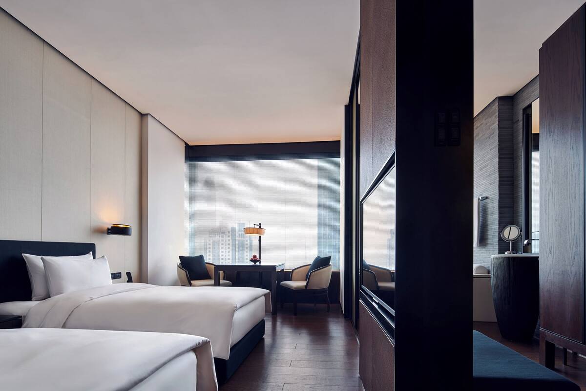 A hotel room with two beds, a desk, a chair, a large window, and a view of the city.