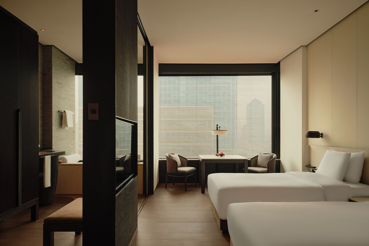 A hotel room with two beds, a desk, a chair, a lamp, and a window with blinds.