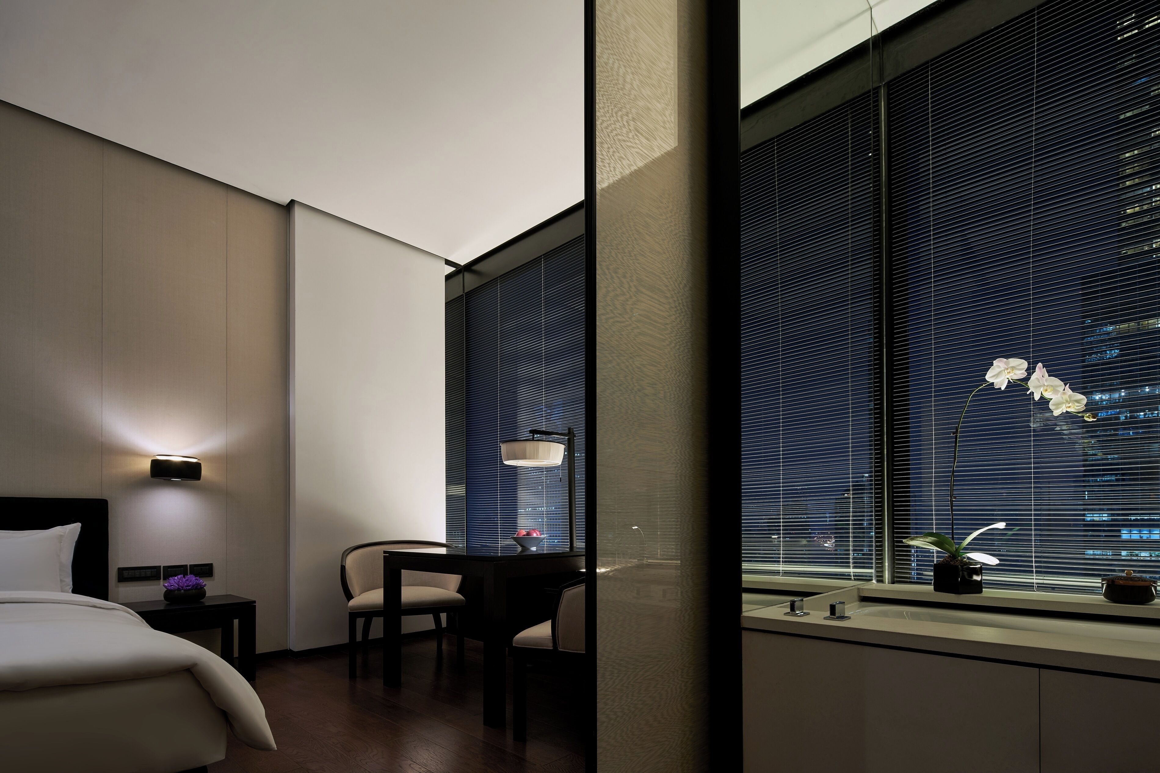 A hotel room with a bed, a desk, a chair, a window with blinds, and a nightstand with a lamp.