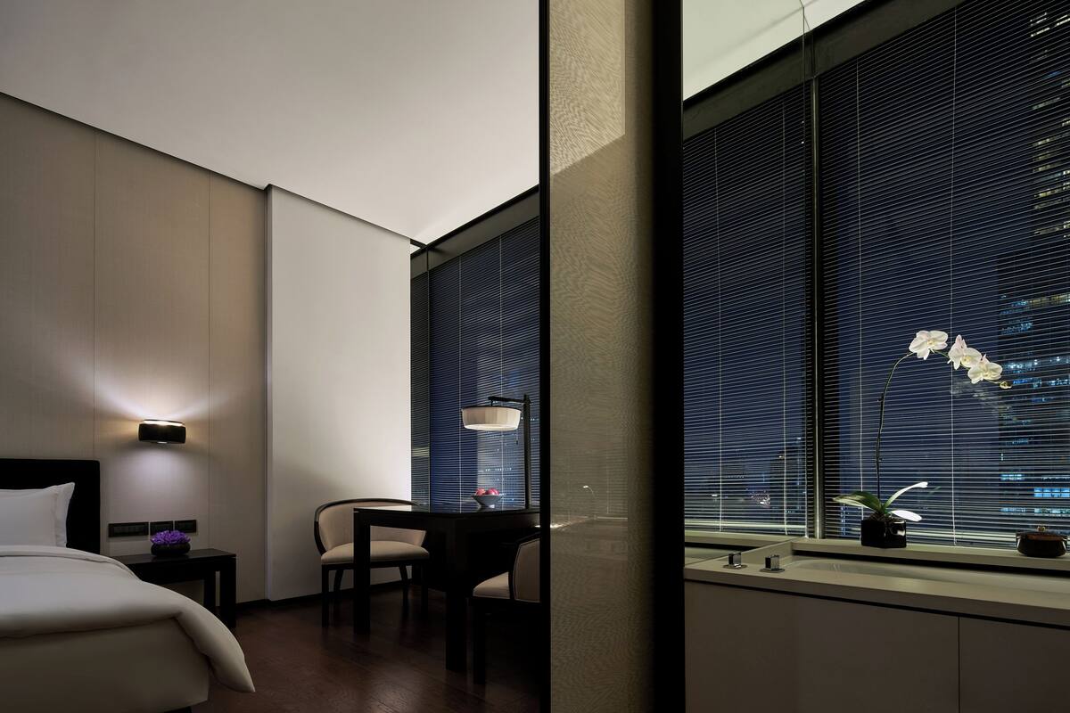 A hotel room with a bed, a desk, a chair, a window with blinds, and a nightstand with a lamp.