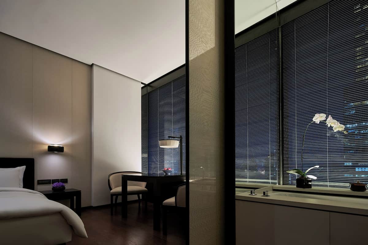 A hotel room with a bed, a desk, a chair, a window with blinds, and a nightstand with a lamp.
