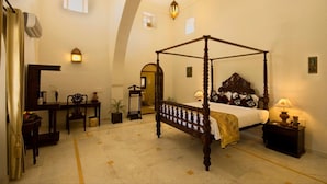 A four-poster bed in a room with a high ceiling, a desk with a chair, a small table with a lamp, and a potted plant. - Fort Khejarla, Jodhpur (Bilara)