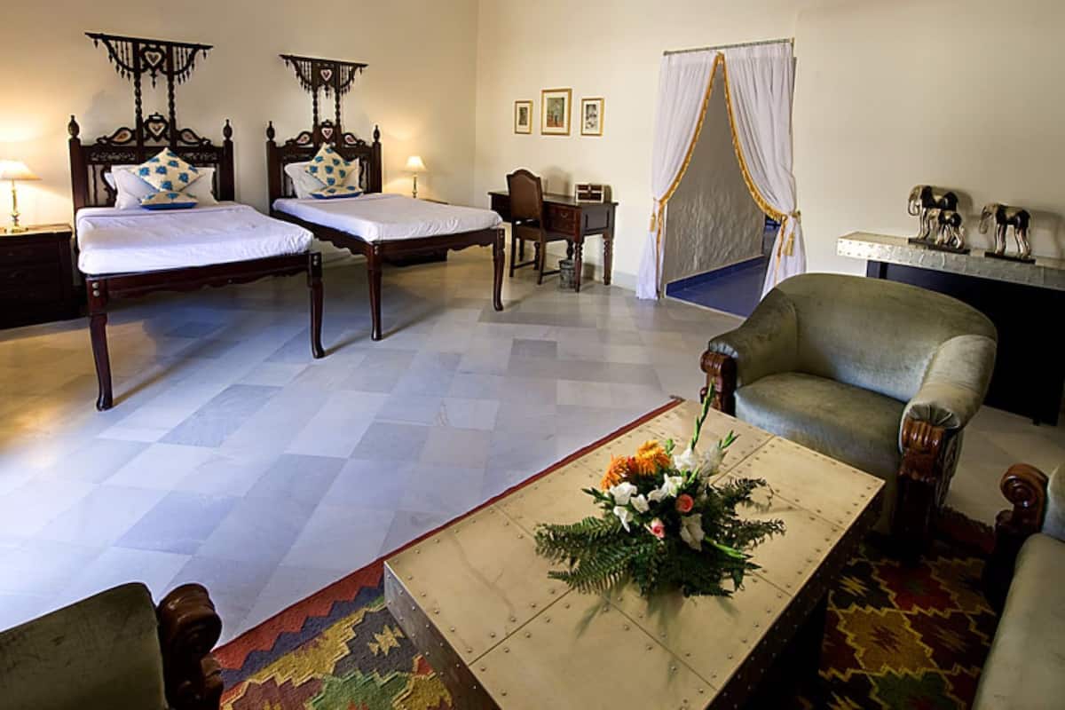 Ein Hotelzimmer mit zwei hölzernen Betten, einem Schreibtisch, einem Stuhl, einem kleinen Tisch mit Blumenarrangement und einer dekorativen Elefantenstatue.