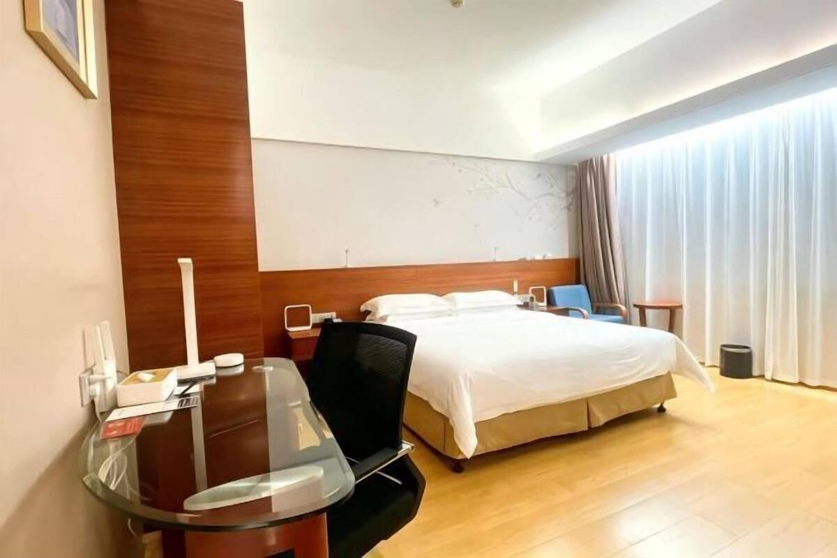 A hotel room with a bed, a glass table, a chair, a bedside table, and a large window with curtains.
