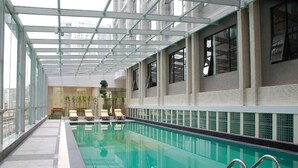 Una piscina interior con paredes de vidrio transparente, suelo de baldosas y algunas sillas de descanso.
