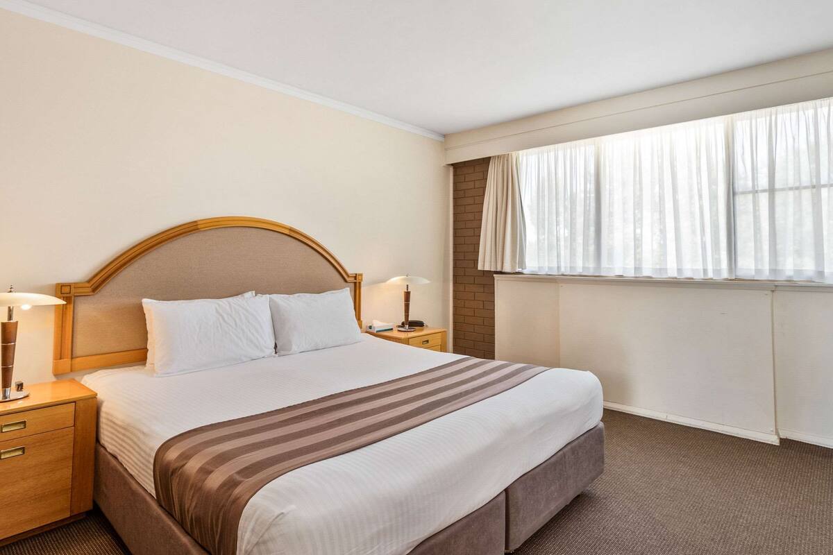 Camera d'albergo con un letto grande, una testiera in legno, due comodini con lampade, una finestra con tende leggere e un termosifone.