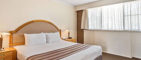 Une chambre d’hôtel avec un grand lit, une tête de lit en bois, deux tables de chevet avec des lampes, une fenêtre avec des rideaux légers et un radiateur.