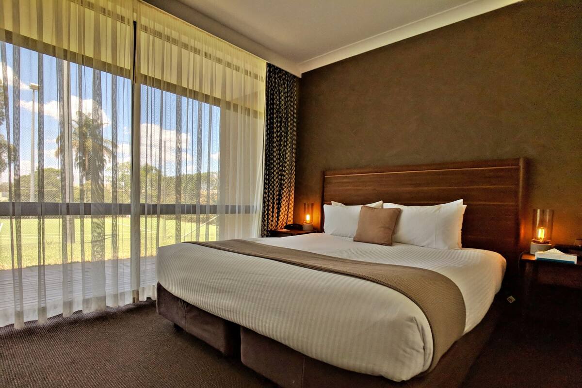 Un letto grande con testiera in legno, vista su un prato verdeggiante attraverso tende trasparenti e un comodino con lampada.
