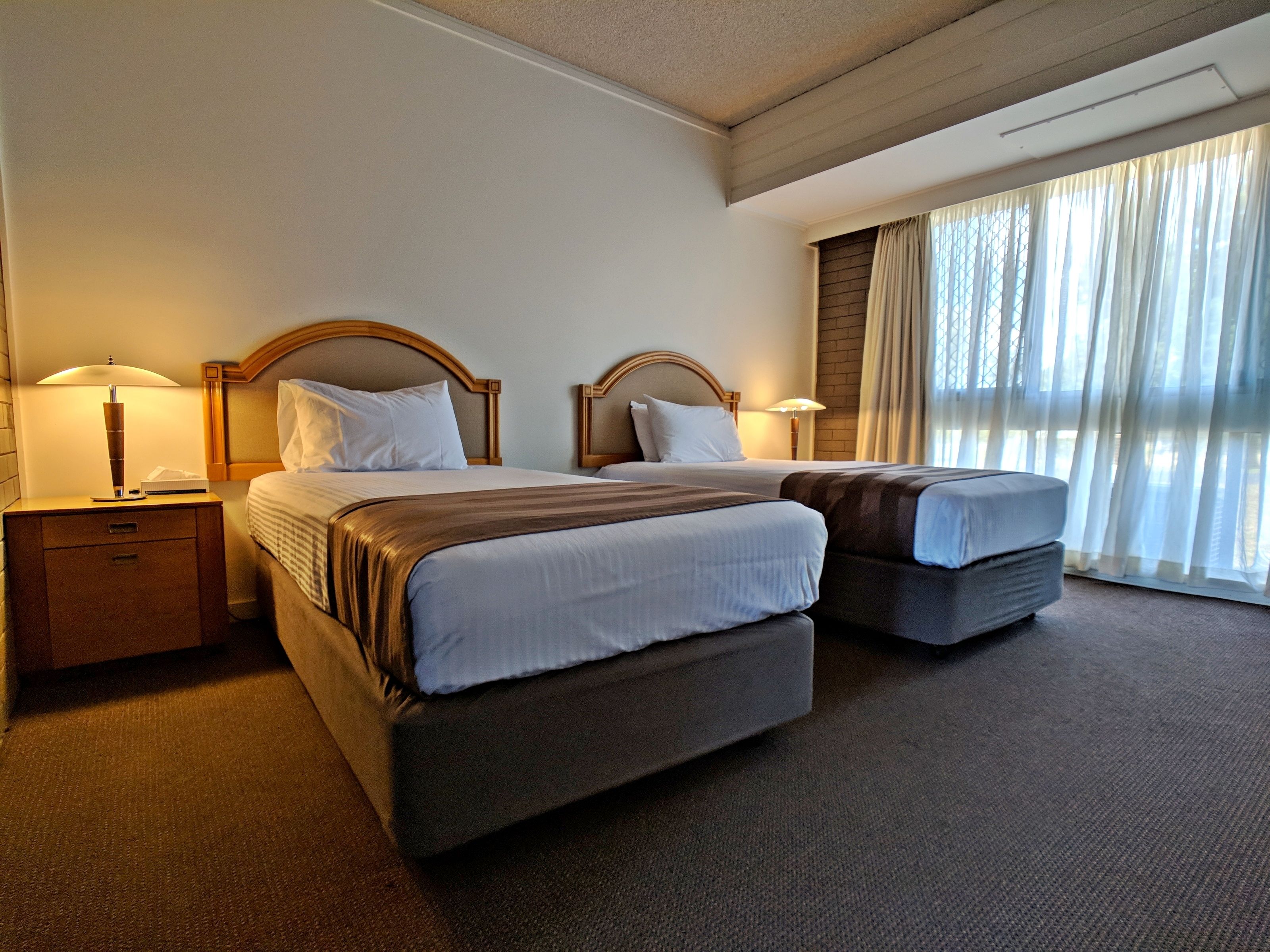 A hotel room with two beds, a nightstand, a lamp, and a large window with curtains.
