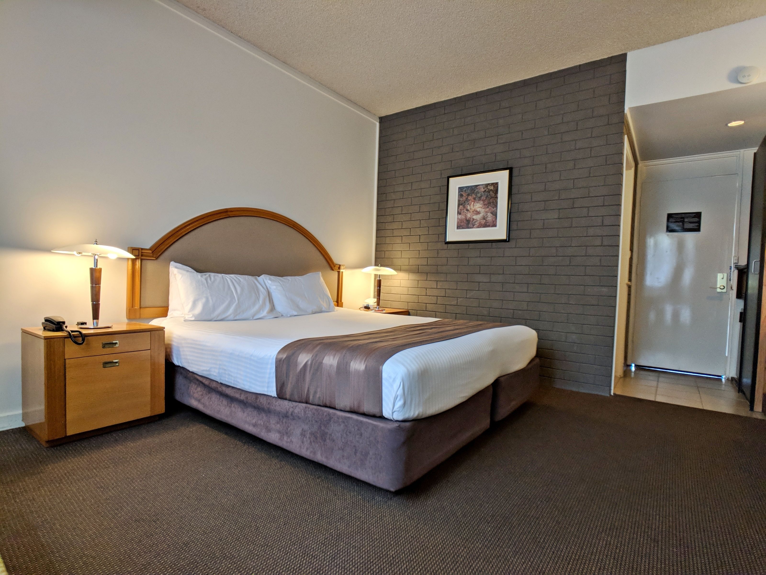 A hotel room with a bed, bedside lamps, a nightstand, a door, and a wall with a framed picture.