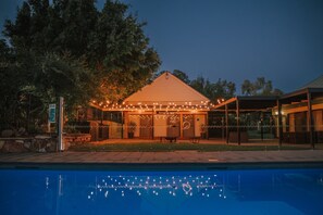 Zona de piscina con un área de descanso cubierta, un cartel y un edificio con luces colgantes.