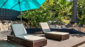 Dos sillones de mimbre con cojines bajo un paraguas turquesa, sobre una terraza de madera con un muro de piedra y vegetación al fondo.