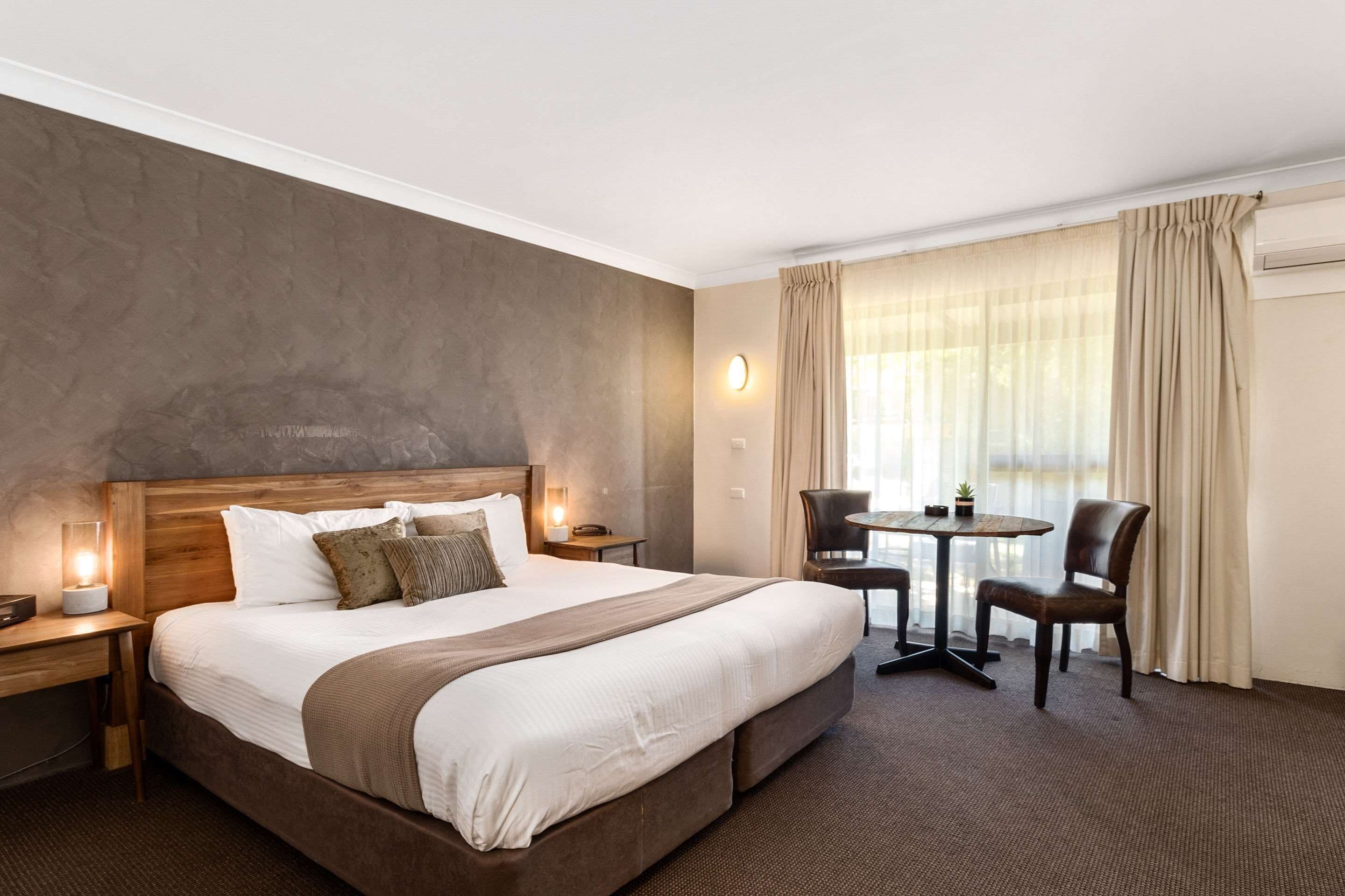 A hotel room with a large bed, bedside tables, a desk, a chair, a small table, and a window with curtains.
