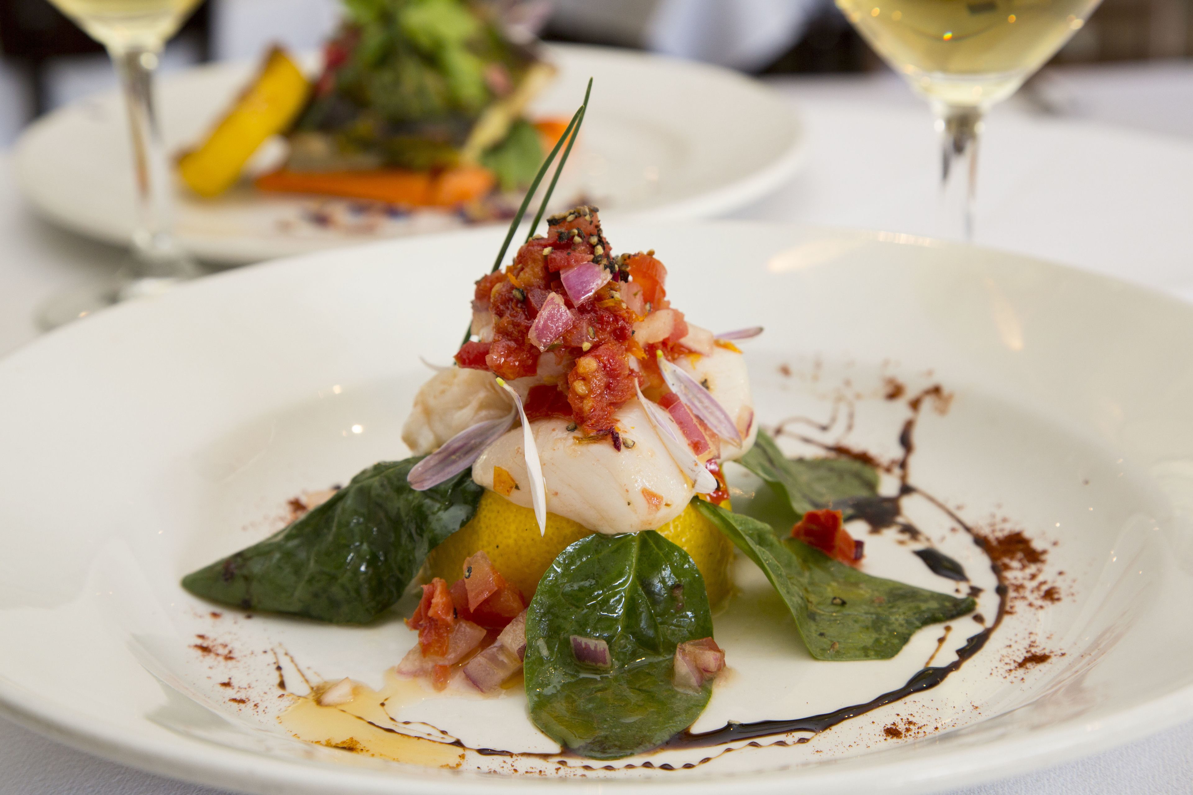 Une assiette blanche contenant un plat de poisson, des légumes verts et des légumes coupés en dés, accompagné d’un verre de vin blanc.