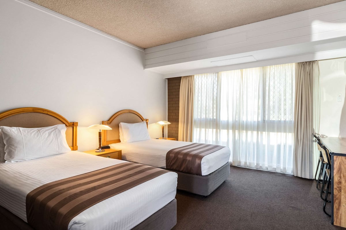 Camera d'albergo con due letti, una scrivania con sedie e un'ampia finestra con tende.