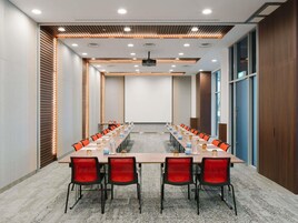 A conference room with a long table, red chairs, a projector screen, and a wooden accent wall.