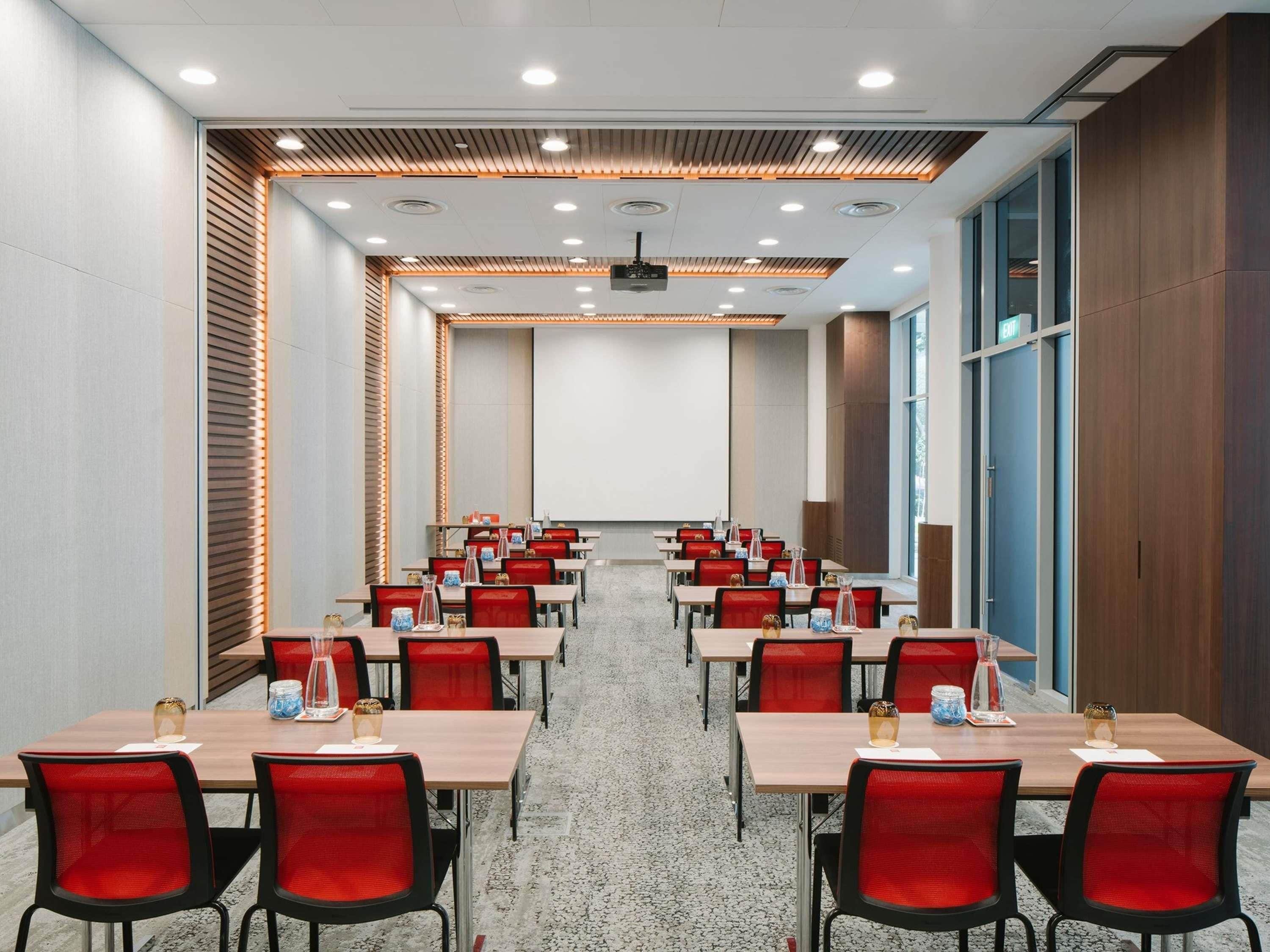 Une salle de conférence équipée de plusieurs tables et chaises, d’un écran de projection et d’une grande fenêtre.