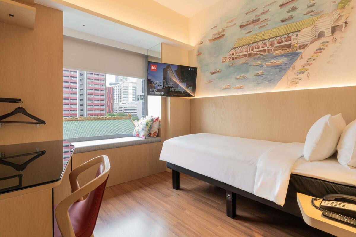 Habitación de hotel con cama, escritorio, silla, televisor montado en la pared y ventana con vistas a edificios.