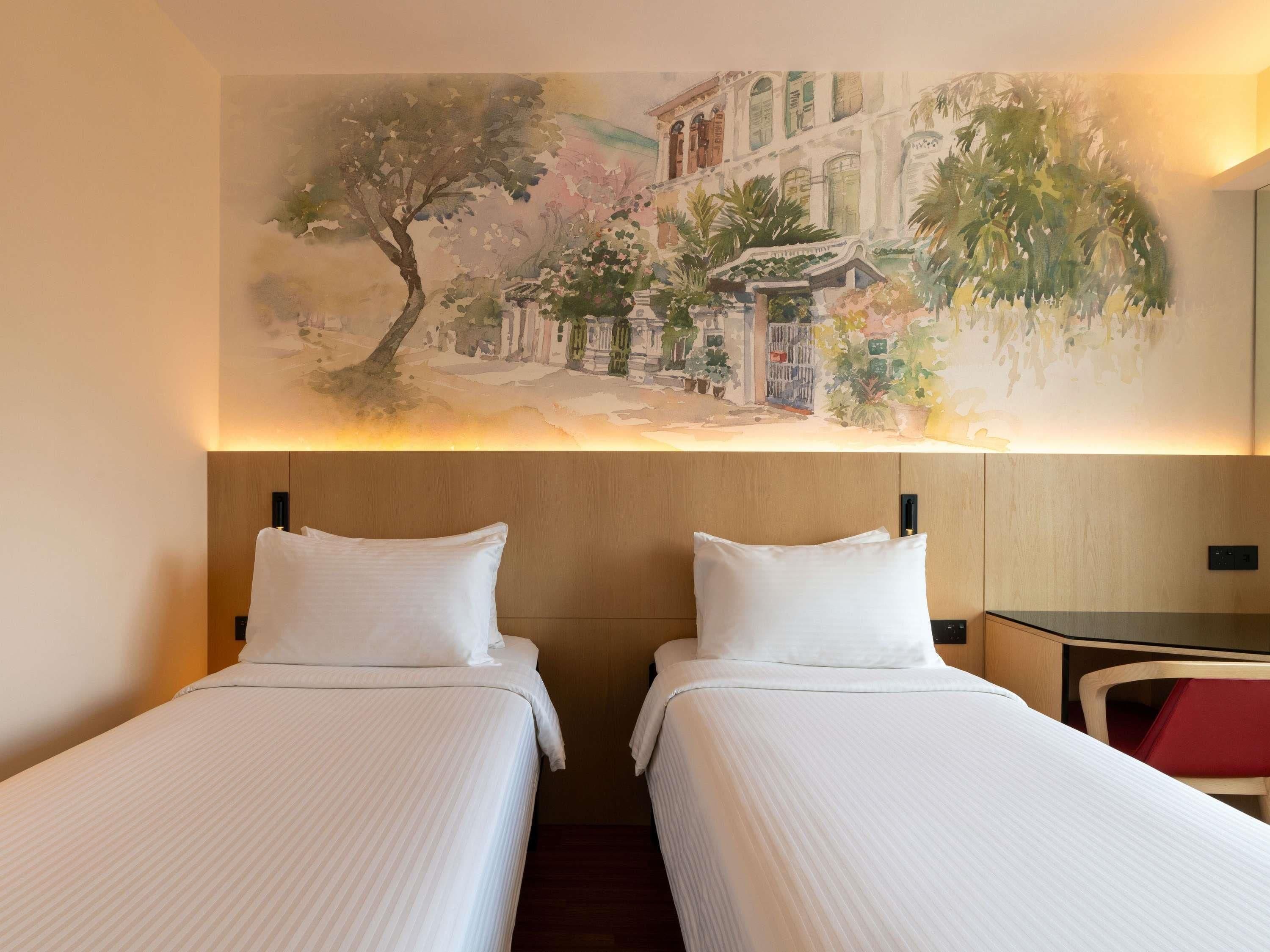 A hotel room with two beds, a mural of a street scene, and a desk with a chair.