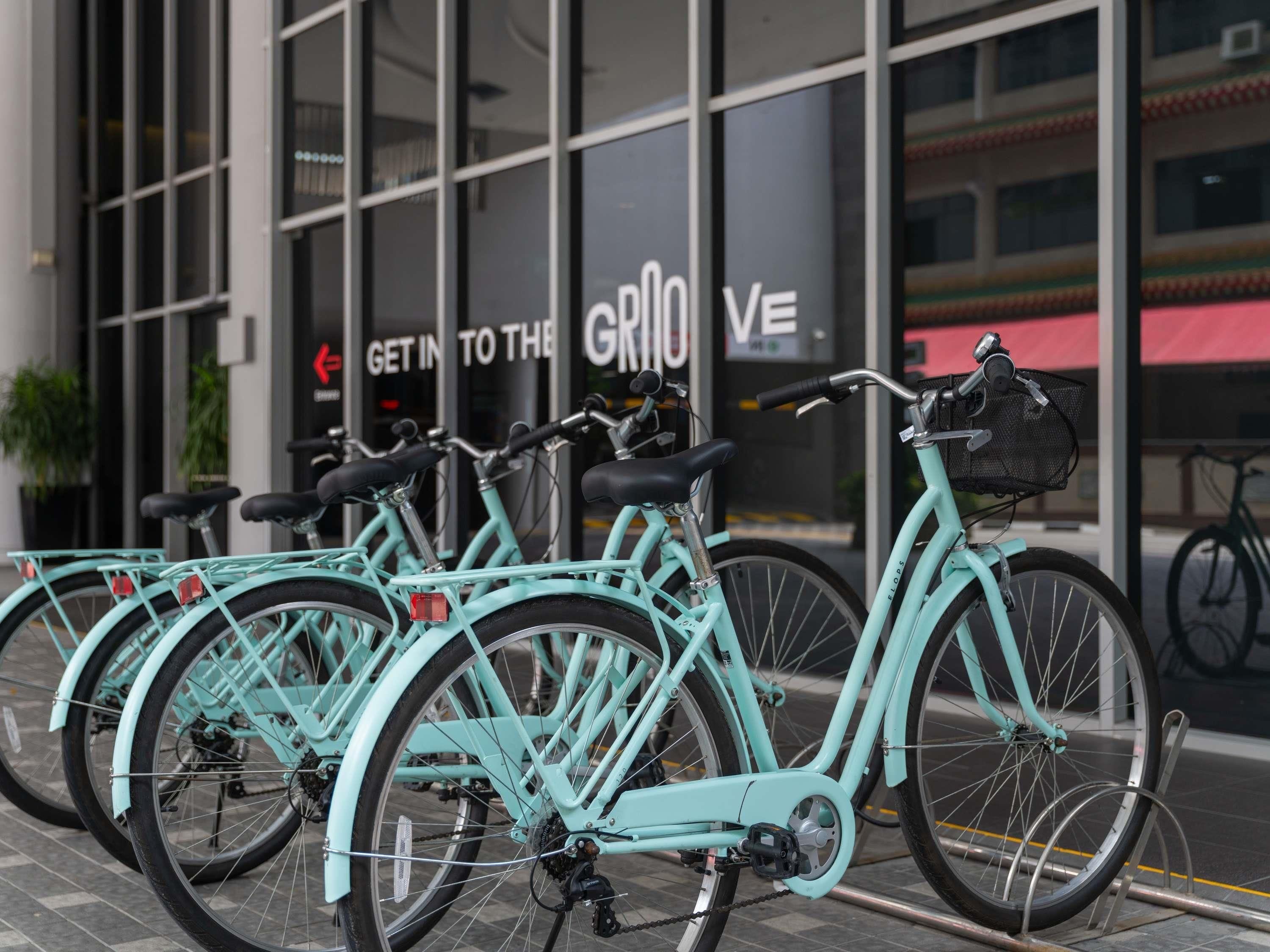 Una fila de bicicletas turquesas estacionadas frente a un edificio con grandes ventanales y un cartel que dice 'Get in to the Grio'.