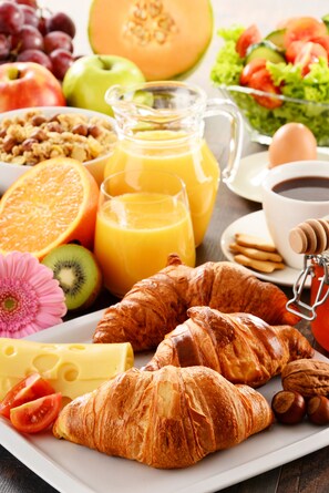 Een ontbijtplateau met croissants, kaas, tomaten en diverse soorten fruit en sap.