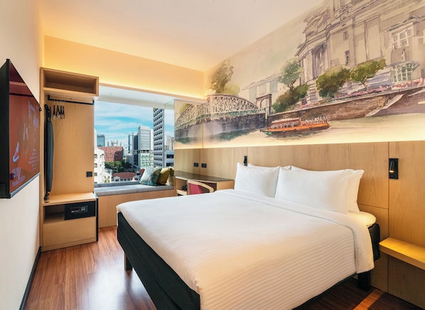 Een hotelkamer met een bed, bureau, kledingkast en een groot raam met uitzicht op de stad.