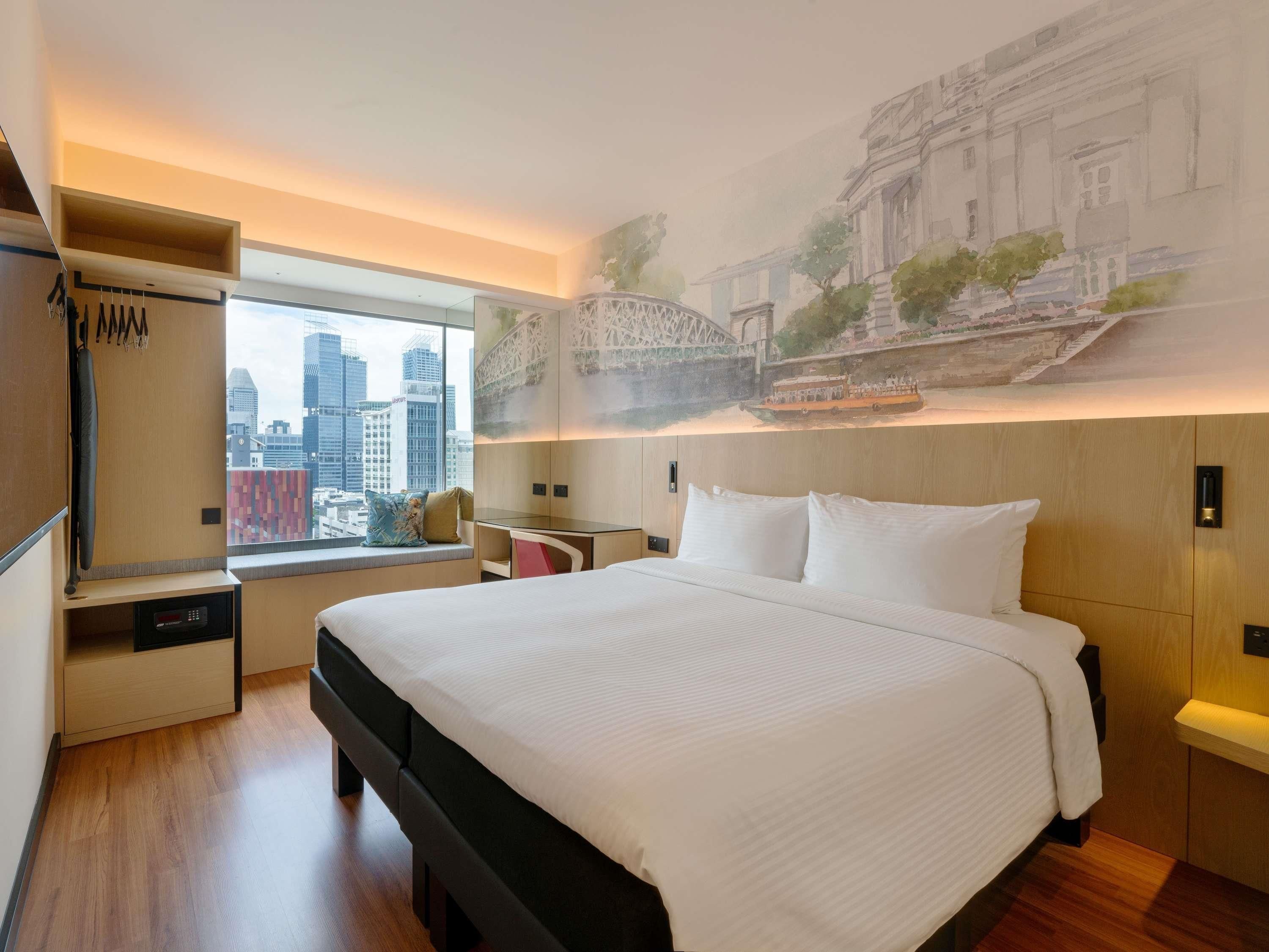 A modern hotel room with a large bed, a city view, a mural of a bridge, and a wardrobe.