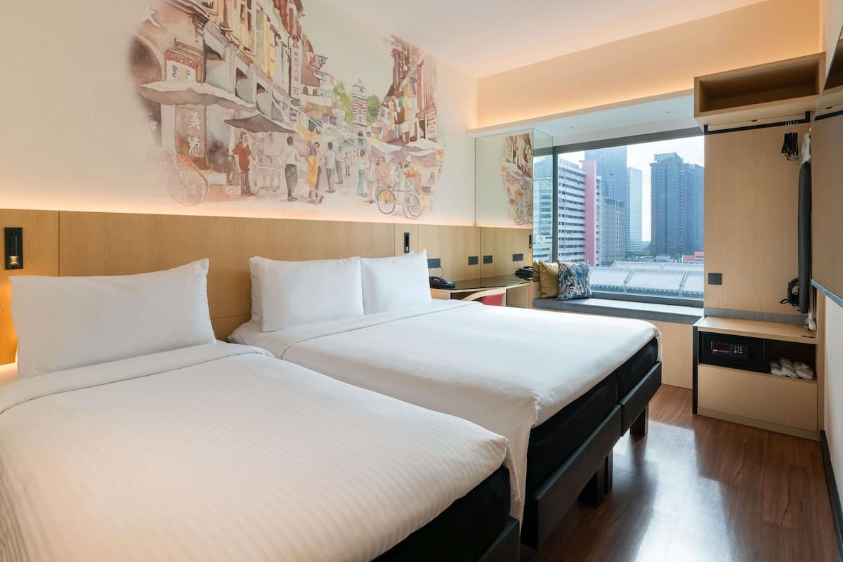 Habitación de hotel con dos camas, un escritorio, una silla, un ventanal con vistas a la ciudad y un televisor de pared.