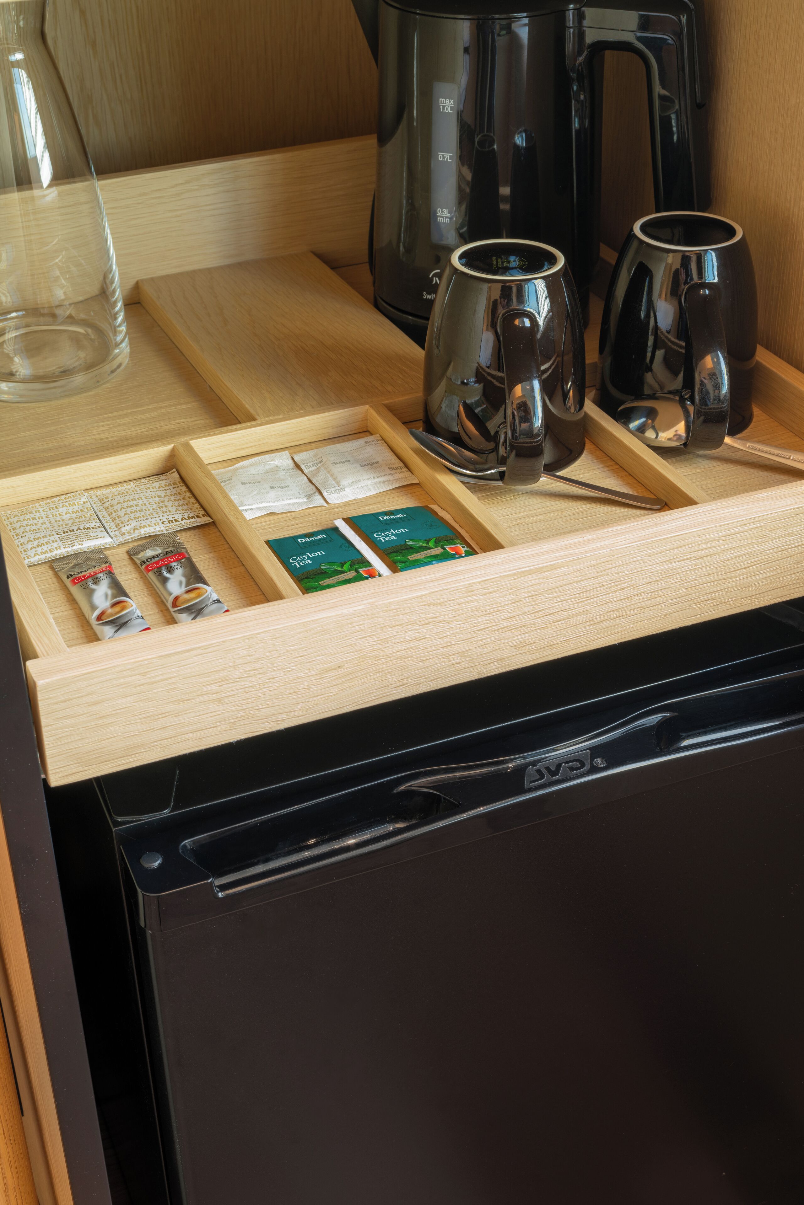 A kitchen counter with a coffee maker, two mugs, a glass, and a drawer with coffee packets and a book.