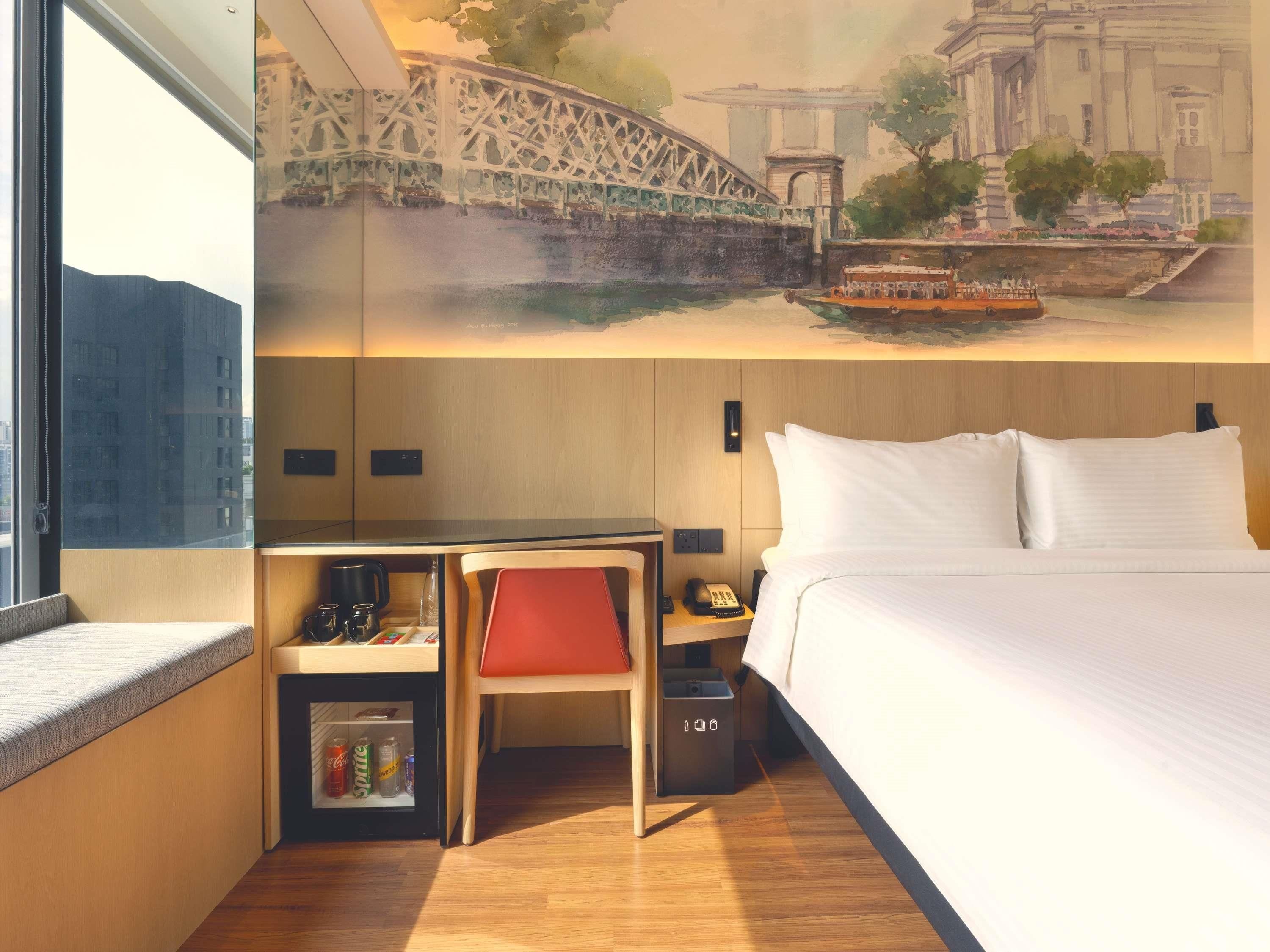 A hotel room with a bed, a desk with a chair, a small cabinet, and a window.