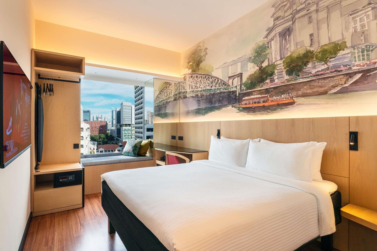 A hotel room with a bed, a seating area with a view, a wardrobe, and a wall mural.