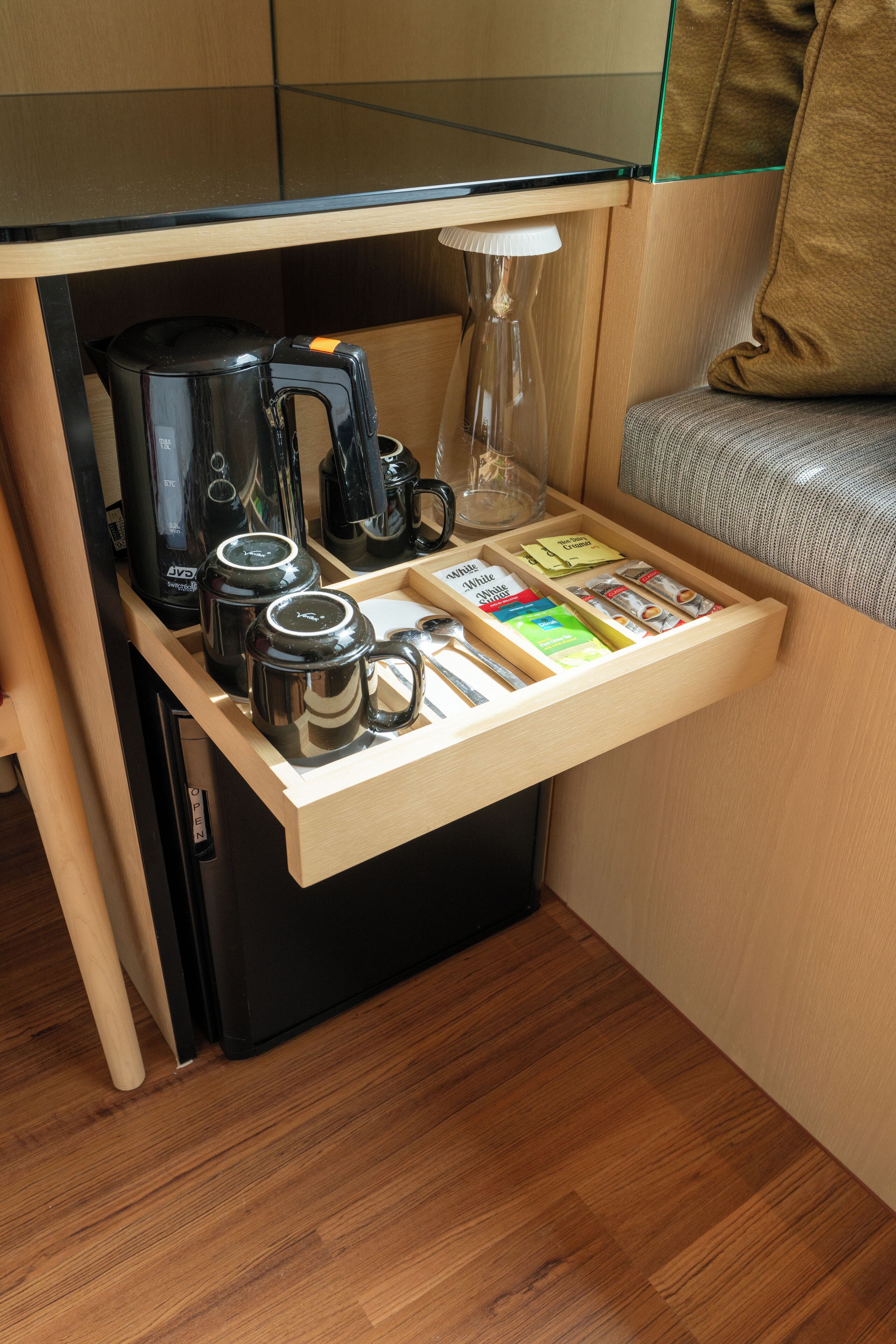 A compact kitchenette with a coffee maker, two mugs, a kettle, and a drawer with snacks and a book.