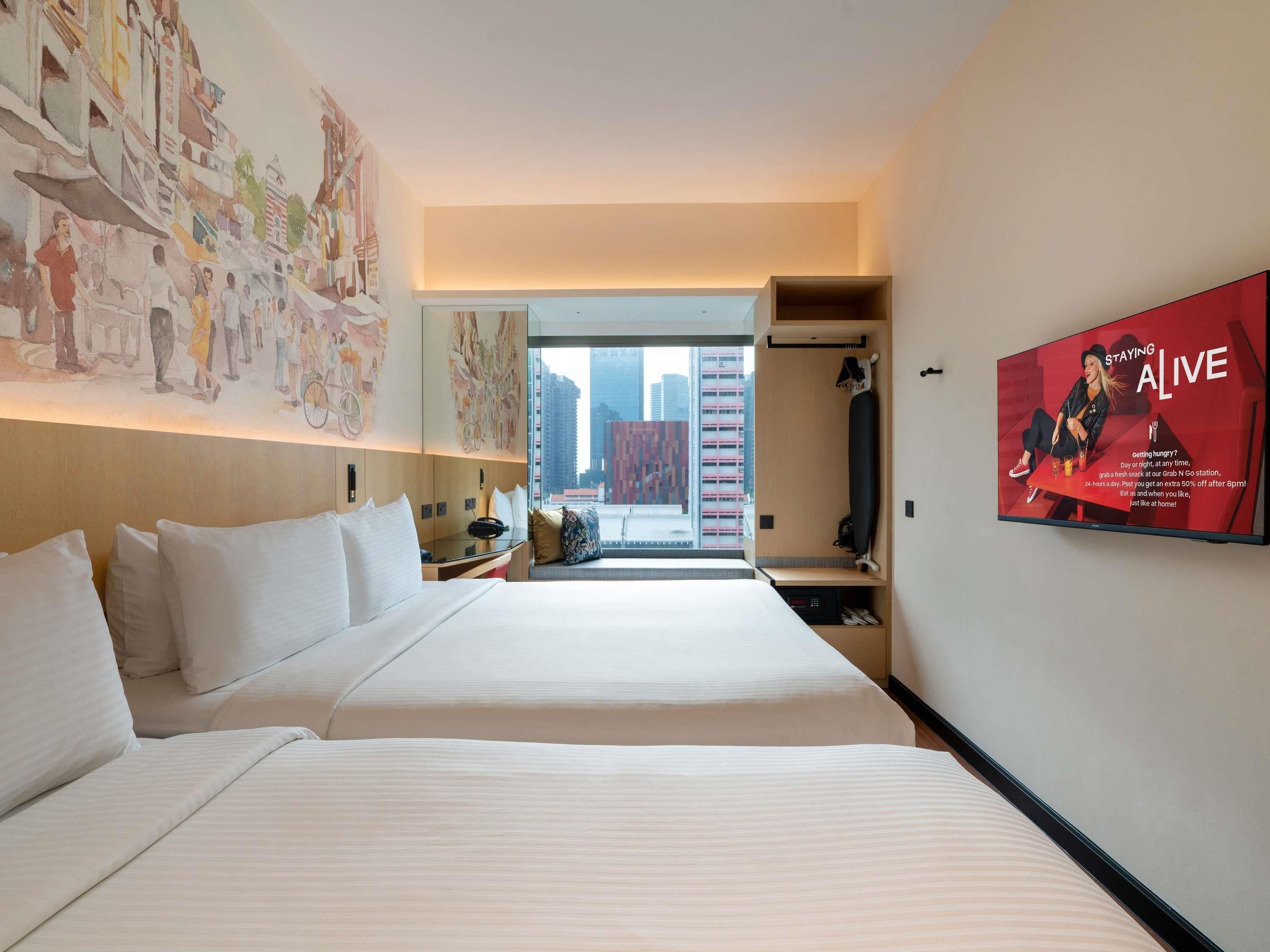 A hotel room with two beds, a large window, a wall-mounted TV, and a view of the city.