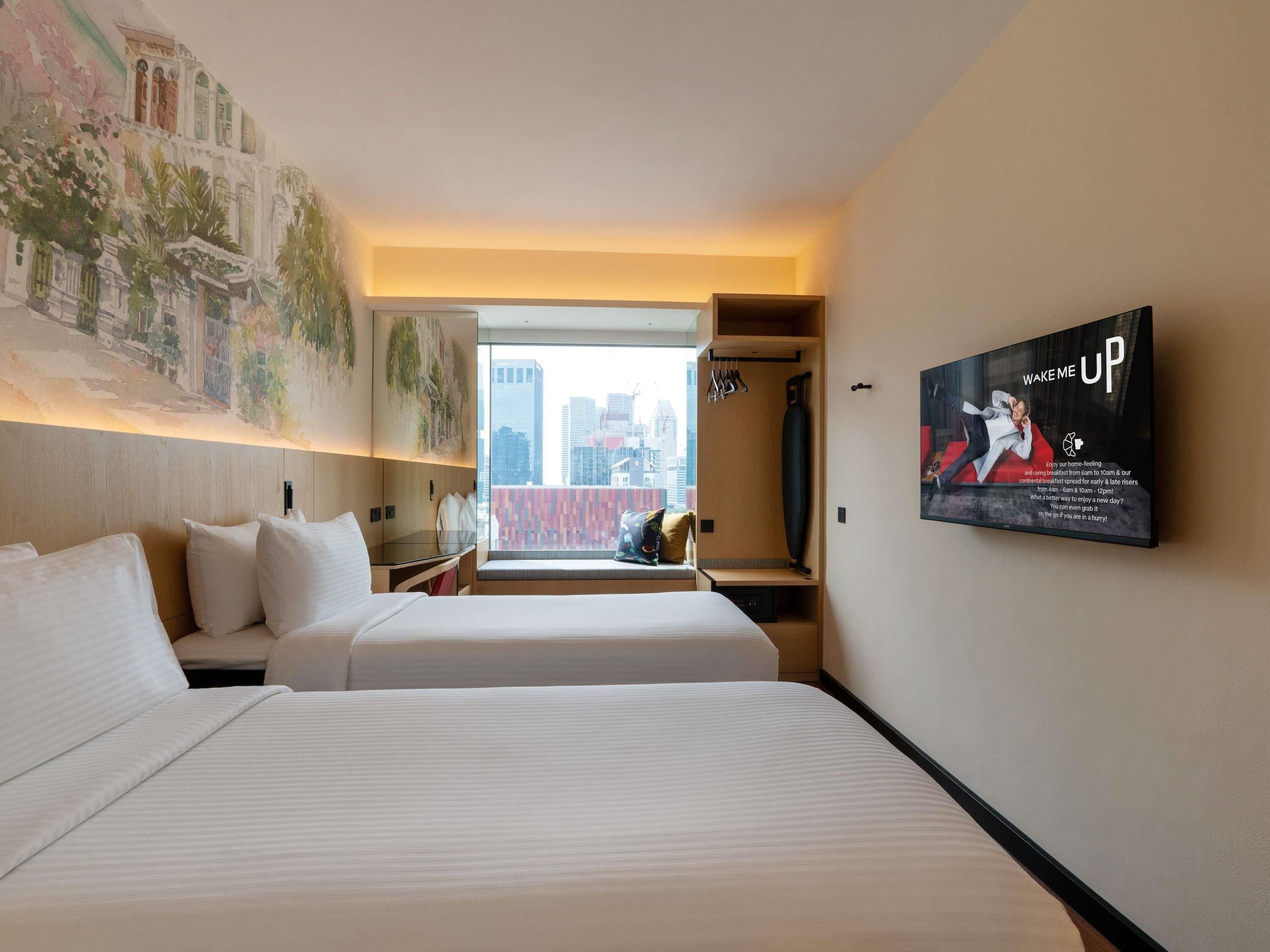 A hotel room with two beds, a large window with a city view, a mounted TV, and a wall with a painting.