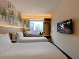 Habitación de hotel con dos camas, un ventanal con vistas a la ciudad, un televisor montado y un cuadro en la pared.