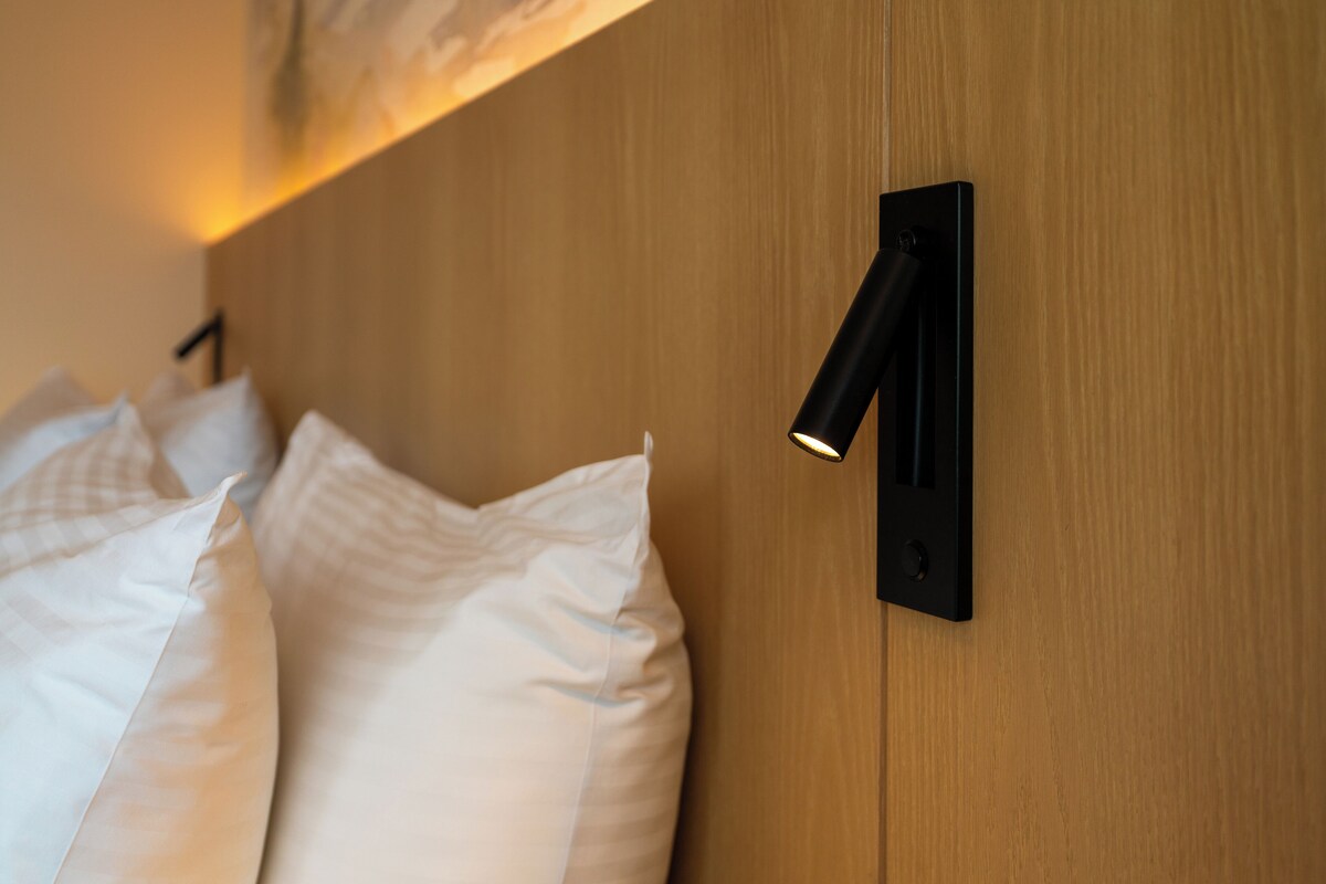 En pænt redt seng med hvide sengetøj og en moderne vægmonteret lampe.