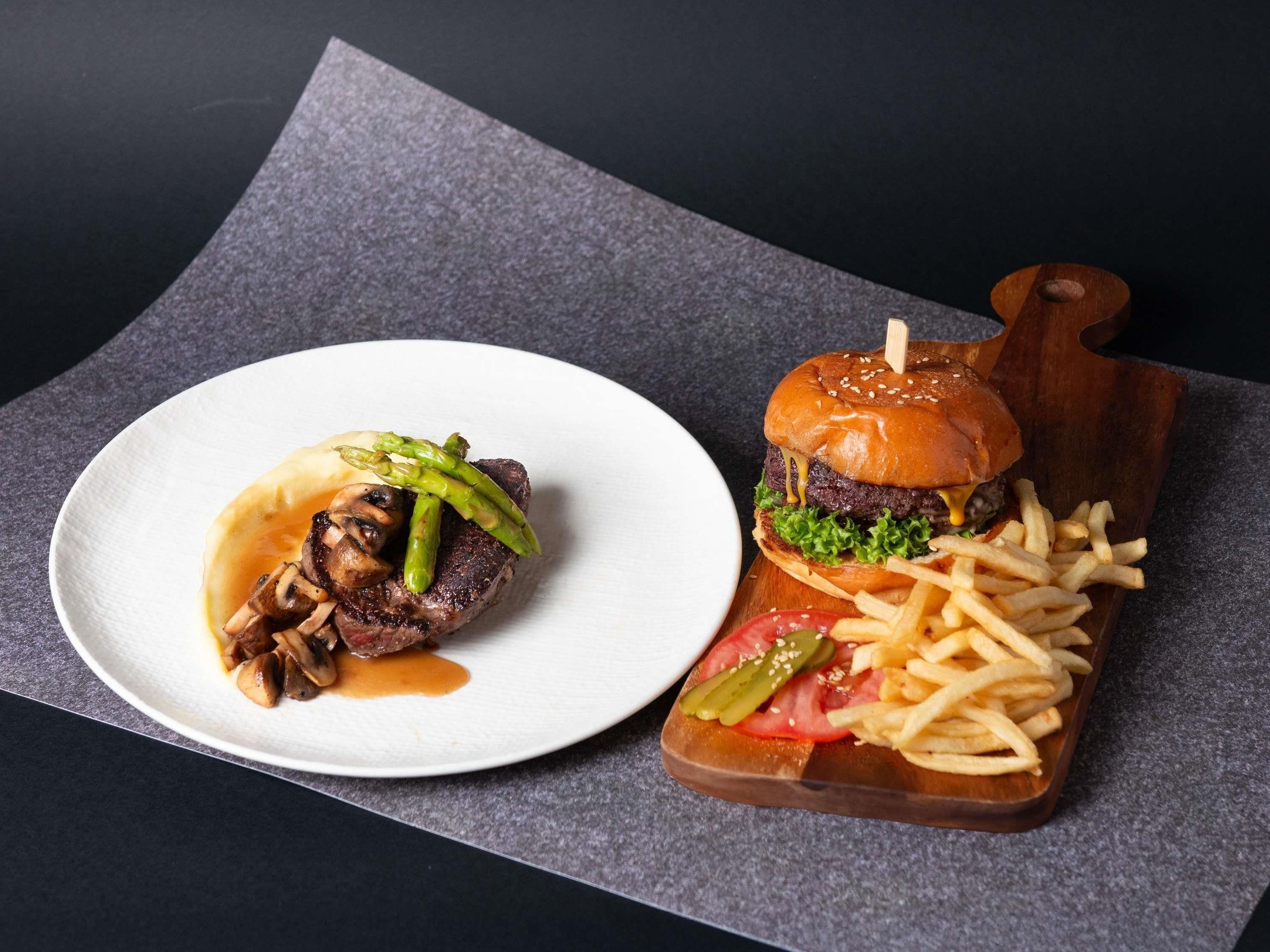 A plate with steak and asparagus, a burger with fries, and a wooden board with a pickle.