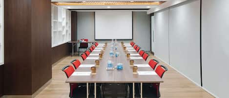 Un salón de conferencias con una larga mesa, sillas, una pantalla de proyector y una pizarra.