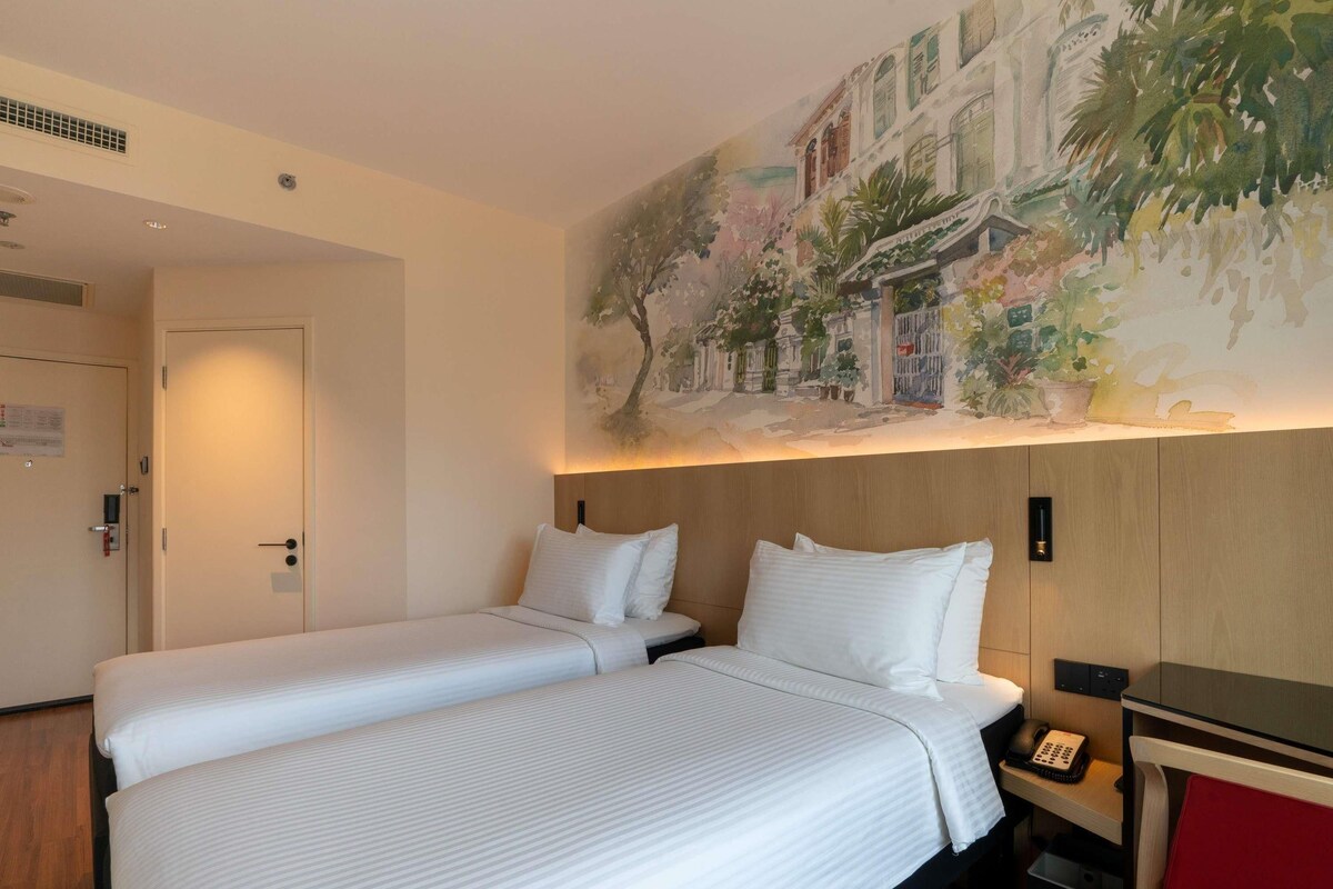 Hotelkamer met twee bedden, een houten hoofdbord, een schilderij van een straatbeeld, een bureau met telefoon en een rode stoel.