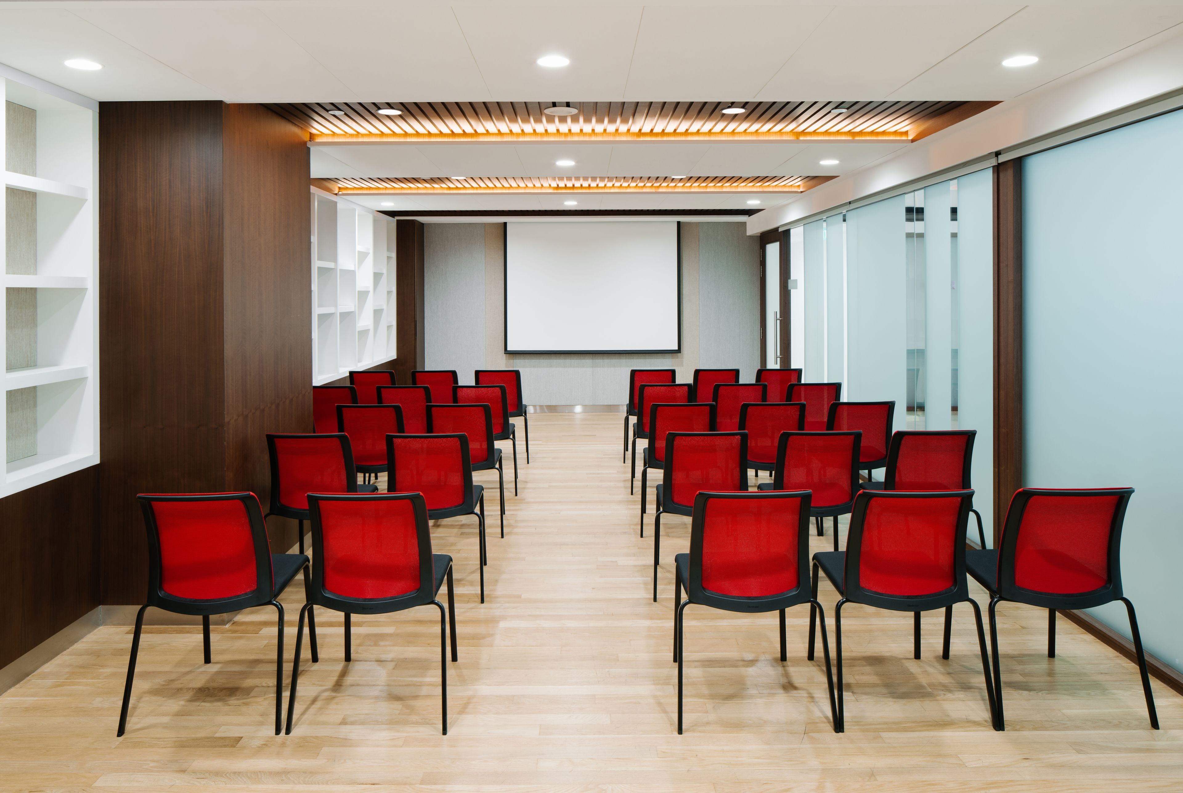 A conference room with rows of red chairs, a projector screen, and a wooden panel wall.