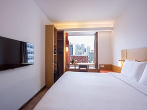 A hotel room with a bed, a TV, a desk with a chair, a window with curtains, and a city view.