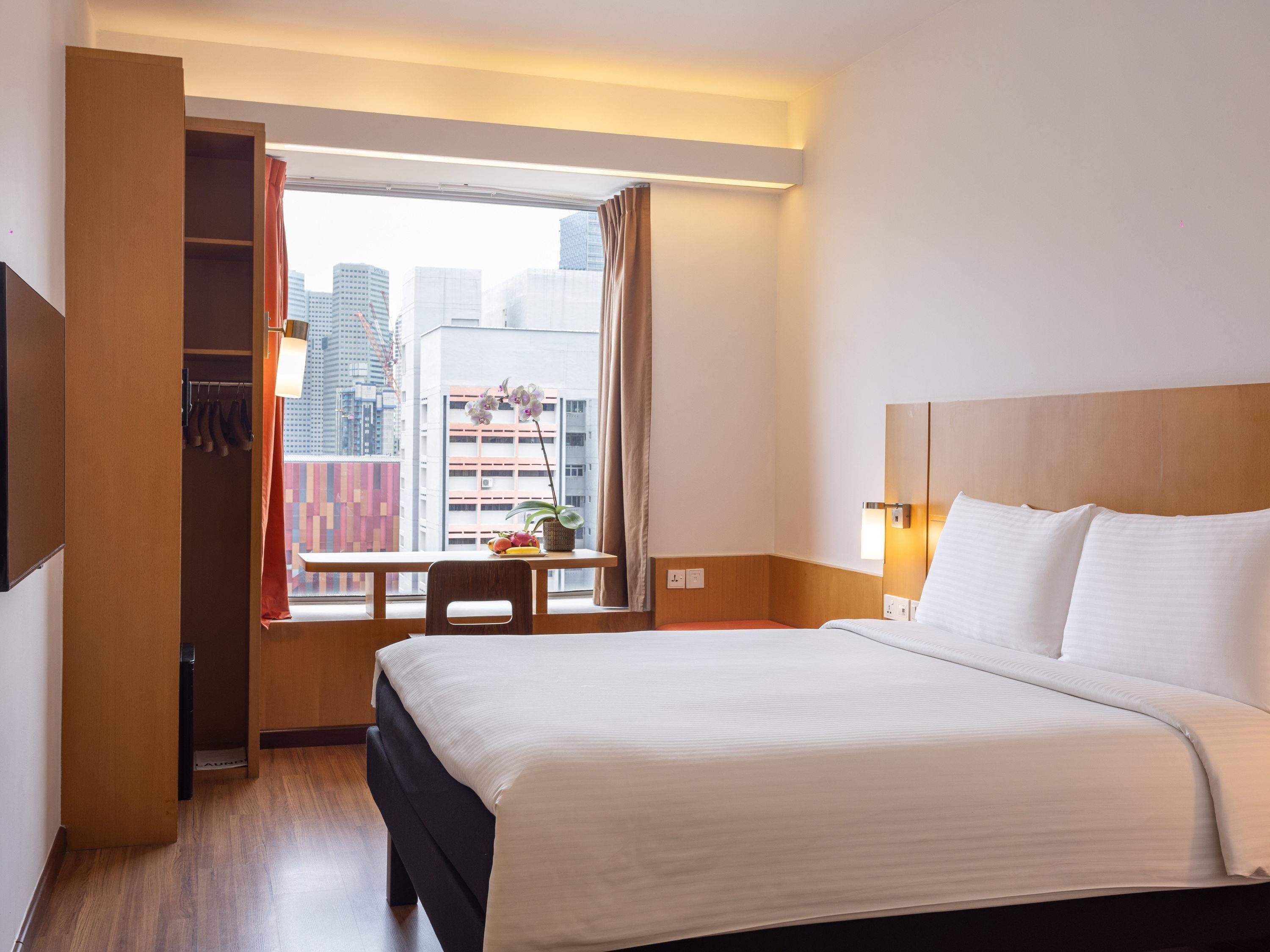 Ein Hotelzimmer mit Bett, einem Fenster mit Vorhängen, einem Schreibtisch mit Pflanze und Blick auf die Stadt.