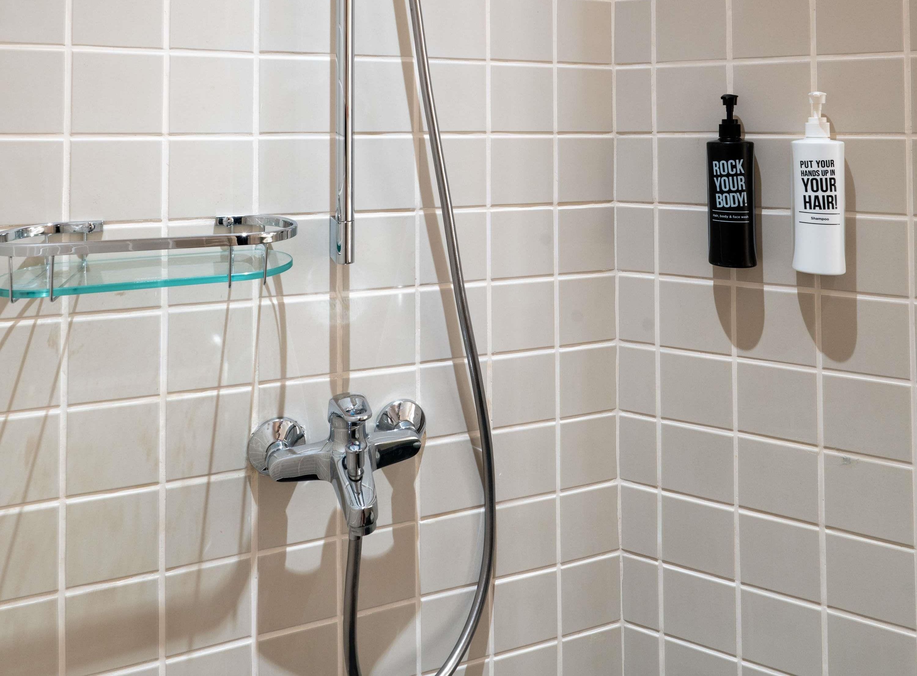 Une salle de bain avec une étagère en verre, une douche dotée d’un robinet chromé et deux bouteilles de shampoing accrochées au mur.