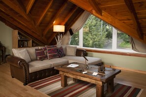 An attic room with a wooden ceiling, a sofa, a coffee table, and a window with a view of trees. - Sol Arrayan Hotel & Spa (Villa La Angostura)